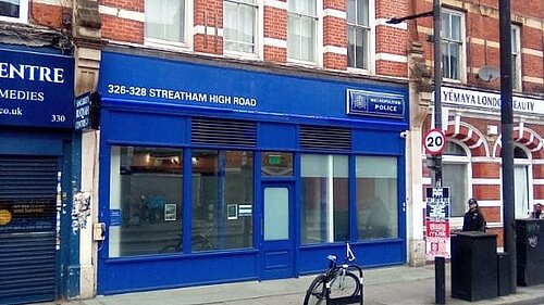 Photo of a road and pavement in the foreground then a line of shops, one of which is a police station